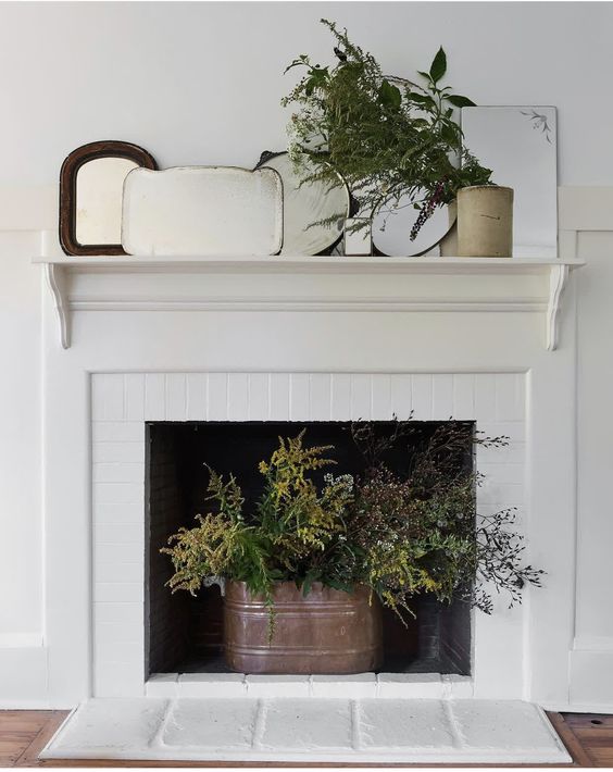 A non working fireplace with a bucket with evergreens, an arrangement of mirrors and greenery on the mantel