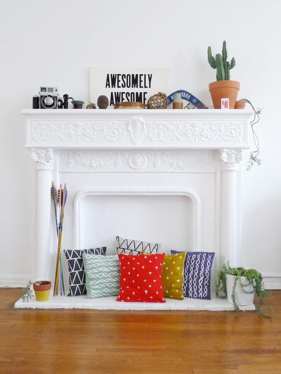 A non working fireplace with an ornated mantel, with colorful pillows, some vintage decor and potted plants