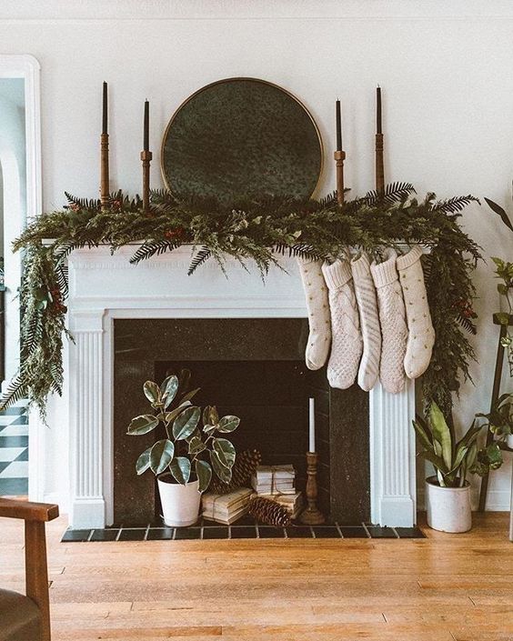 A non working fireplace with books, pinecones, a potted plant and a candle, evergreens, candles and stockings