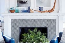 a seaside living room with bold blue chairs, a fireplace with chevron wallpaper, a large seashell with greenery, driftwood, artworks and bottles