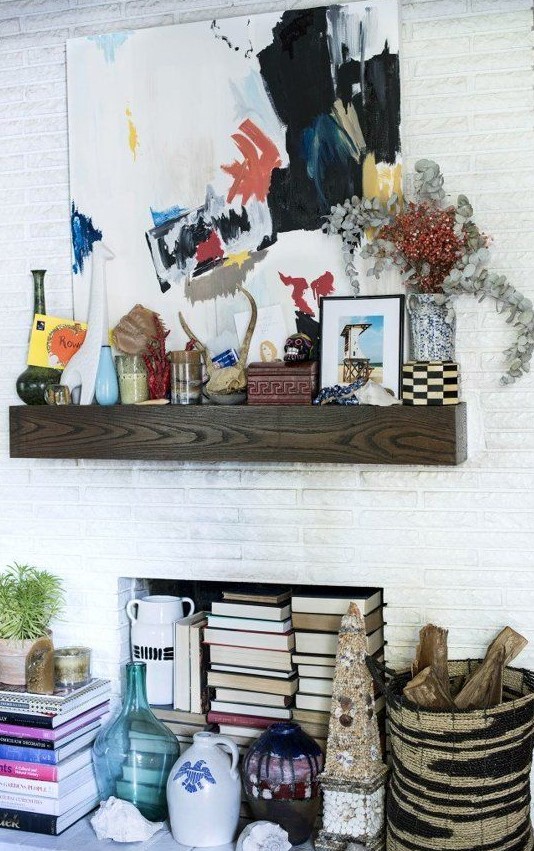 a white brick built-in fireplace with a wood slab mantel, with lots of books inside and candles and various art on the mantel is a stylish idea