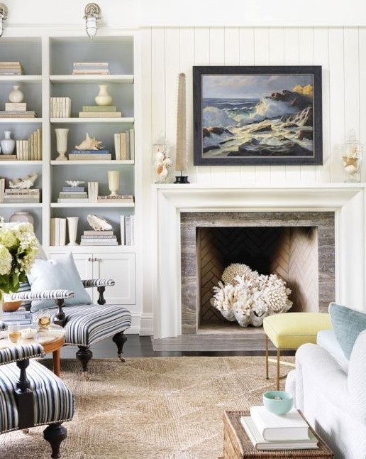 A white coastal living room with built in shelves and a storage unit, a fireplace with a giant seashell and corals, light blue and striped furniture