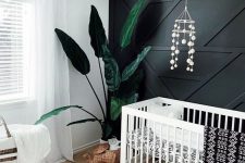 a monochromatic nursery with a black paneled wall, white furniture, a wicker pendant lamp and ottoman and a statement plant