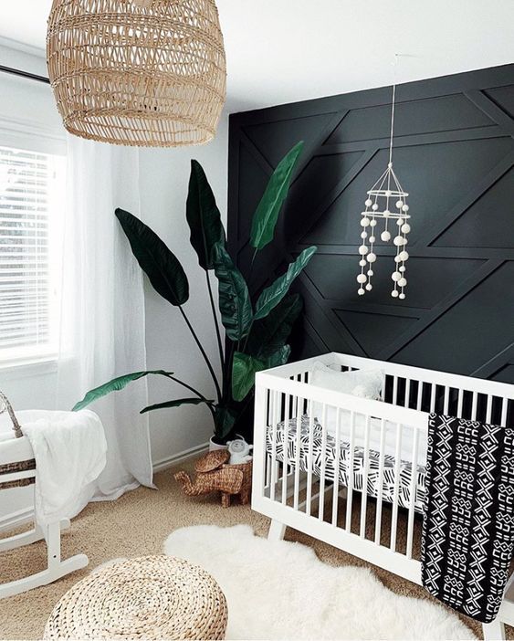 a monochromatic nursery with a black paneled wall, white furniture, a wicker pendant lamp and ottoman and a statement plant
