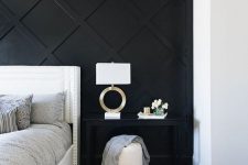 a refined monochromatic bedroom with a black geometric paneled wall, a white bed, a black nightstand, a white pouf and a chic lamp