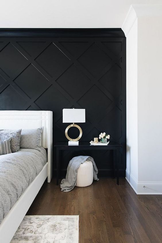 a refined monochromatic bedroom with a black geometric paneled wall, a white bed, a black nightstand, a white pouf and a chic lamp