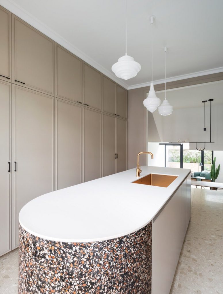 The kitchen is done with tan sleek cabinets that are built-in and are fully closed, while a large curved kitchen island makes a statement with its terrazzo part