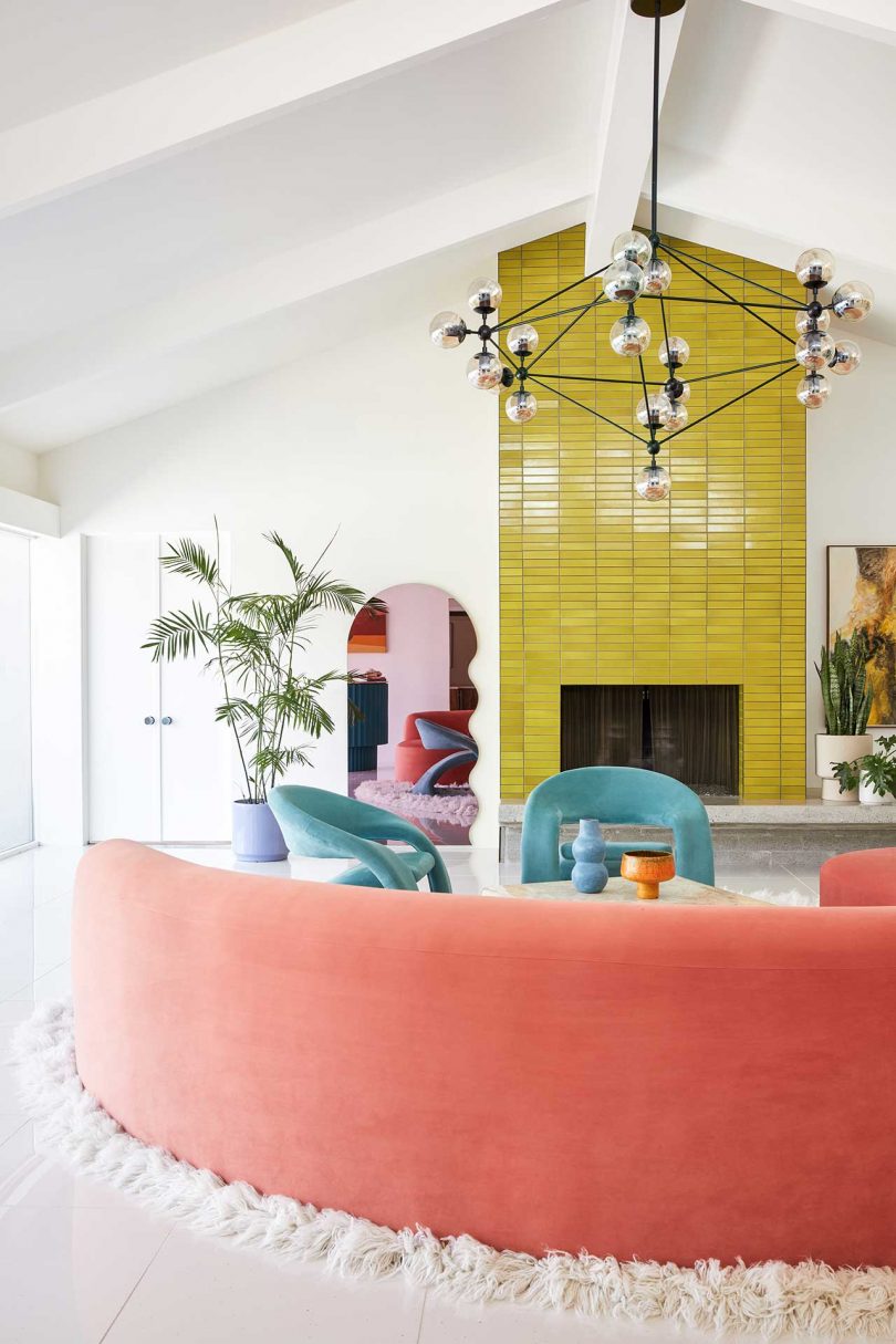 The living room shows off a lemon yellow tile fireplace, some bright furniture, a mid-century modern chandelier and potted plants