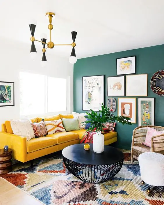 A bright living room with a green wall, a bright yellow sofa, a colorful printed rug, a bright gallery wall and a mid century modern chandelier