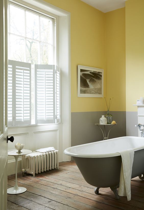 a chic bathroom with grey and yellow color block walls, a grey tub, a wooden floor and shutters for privacy