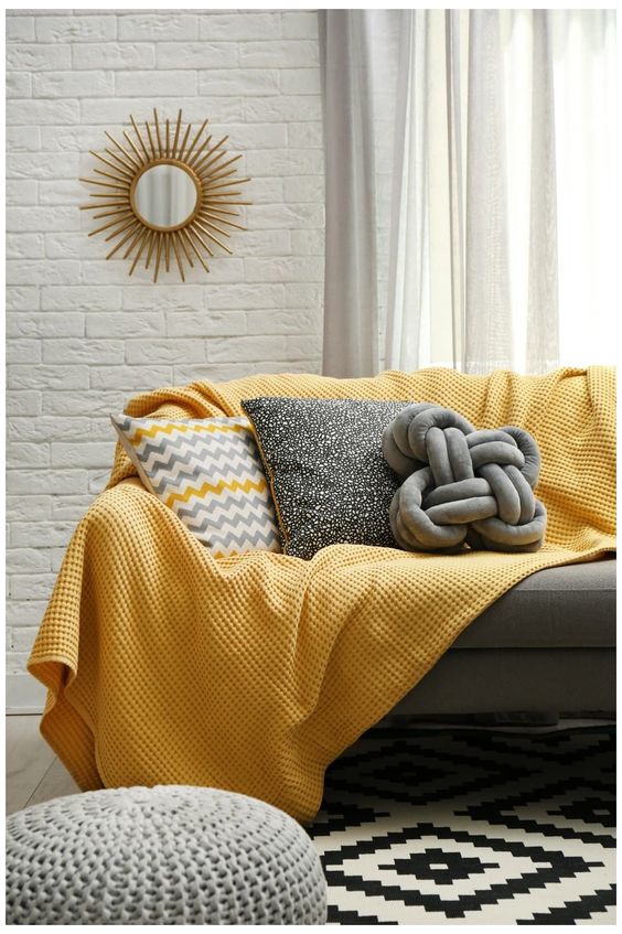 a chic living room with faux white brick walls, a grey sofa, grey and yellow pillows, a grey ottoman and a yellow blanket