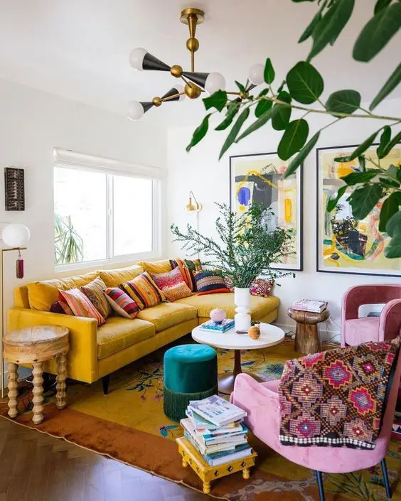 a colorful boho living room with a yellow sofa and a rug, pink chairs, bold printed textiles, a green ottoman, wooden stools and tables