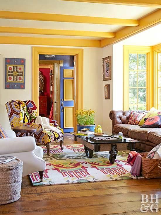 a colorful living room with yellow beams and frames, a brown leather sofa, colorful chairs and a rug, a coffee table on casters