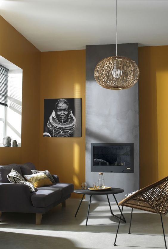 a contemporary living room with mustard walls, a fireplace built into a concrete panel, a grey sofa, a rattan chair and a rattan pendant lamp