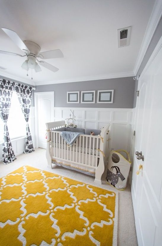 a fun and whimsical nursery with grey and white paneled walls, neutral furniture, grey bedding, a mustard rug and printed curtains