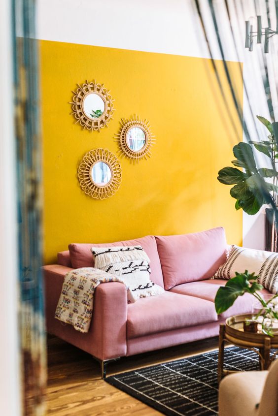 a fun living room with a yellow accent wall, a pink sofa, printed textiles, sunburst mirrors and potted plants
