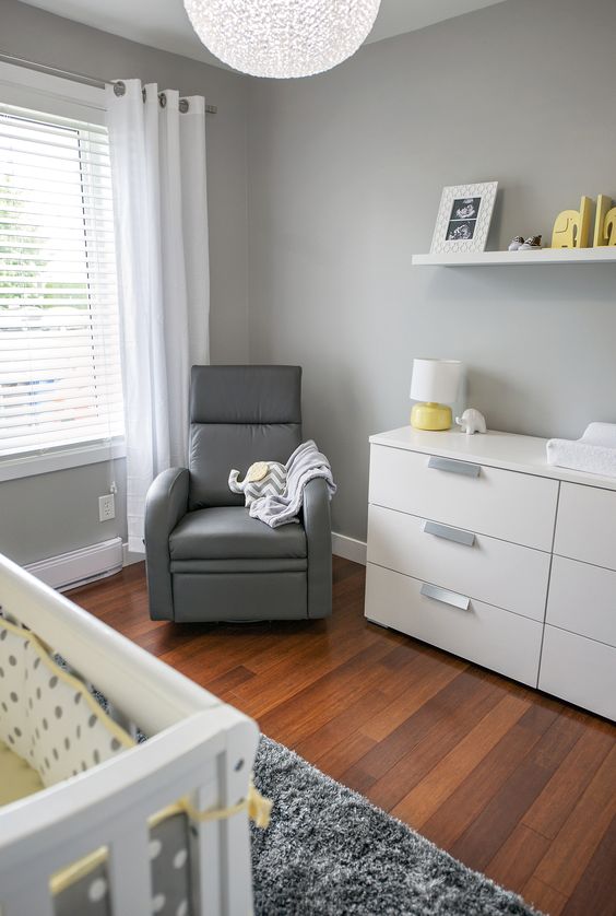 a grey nursery with white furniture, a grey chair, some white, grey and yellow textiles and an open shelf with artworks
