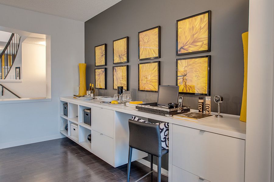 a modern home office with a grey accent wall, a yellow gallery wall of floral prints, a large desk with storage and a grey chair
