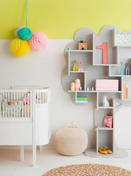 a nursery with color block white and yellow walls, a whisical grey shelf, a white crib and some bold paper pompoms