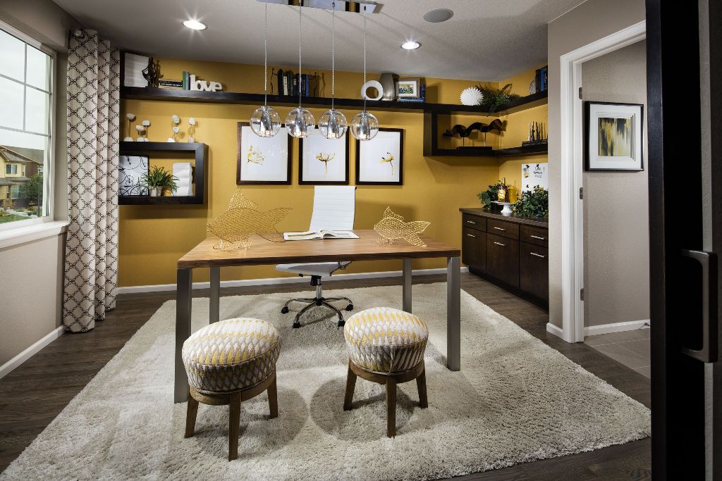 a quirky home office with a honey yellow accent wall and lots of dark open shelves, a gallery wall, a desk, printed grey and yellow ottomans and yellow wire fish