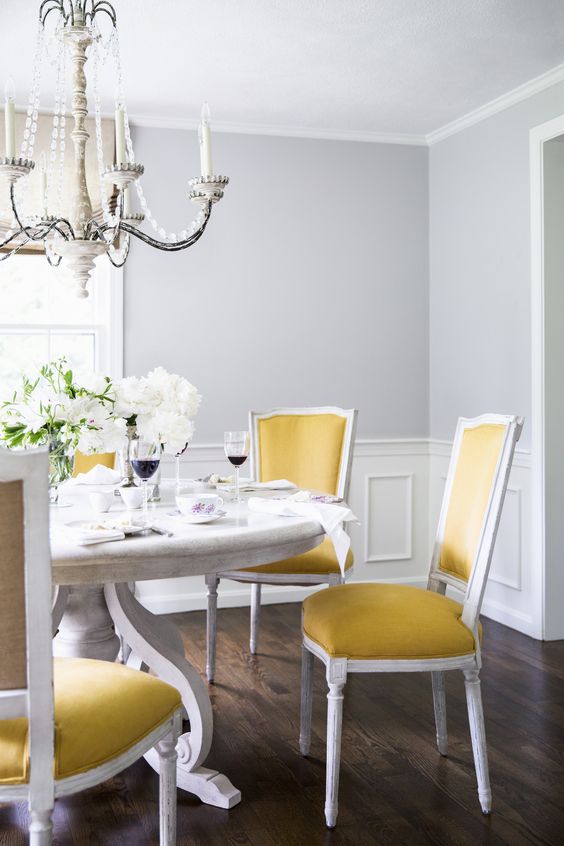 a refined classic dining room with grey walls, paneling, a large white table, yellow chairs, a beautiful crystal chandelier and white blooms