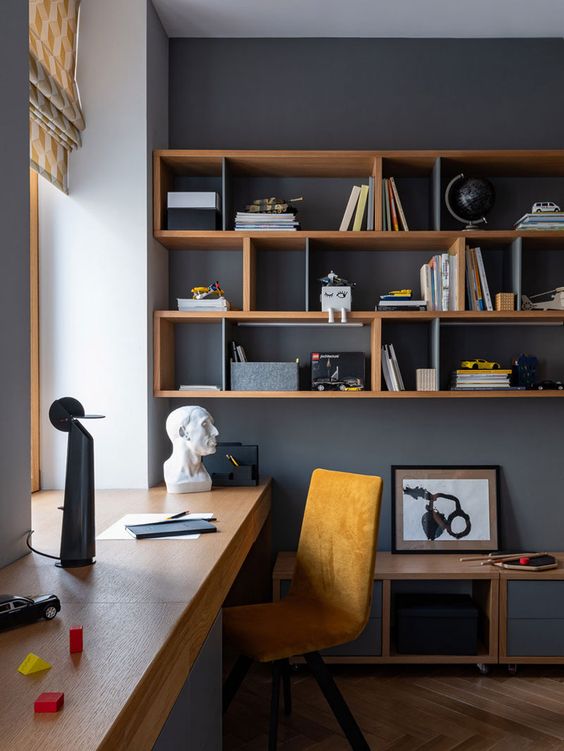 a stylish modern home office with graphite grey walls, a windowsill desk, an open shelving unit, a yellow suede chair and a cabinet