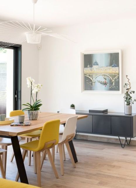 an airy modern dining room with a grey storage unit, a dining table, yellow and white chairs and a pretty retro chandelier