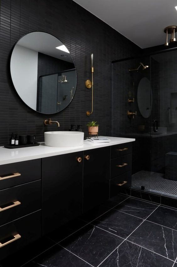 a stylish black bathroom with skinny and marble tiles, a large vanity, a round mirror and brass touches