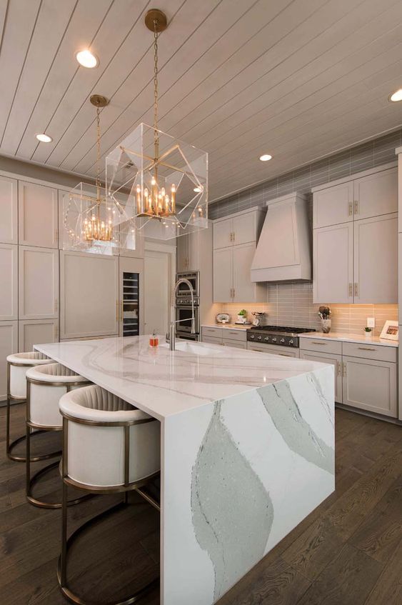 A refined neutral colored kitchen with a jaw dropping kitchen island with a waterfall countertop and stunning gold and sheer glass chandeliers