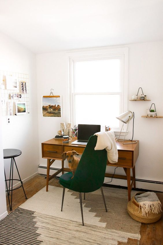 A pretty and cozy mid century modern home office with a vintage desk, a hunter green chair, a basket, some shelves and a grid