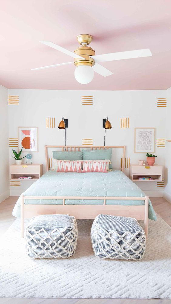 a pretty bedroom with a pink ceiling, light-stained furniture, an accent wall, mint bedding and ottomans in blue and white