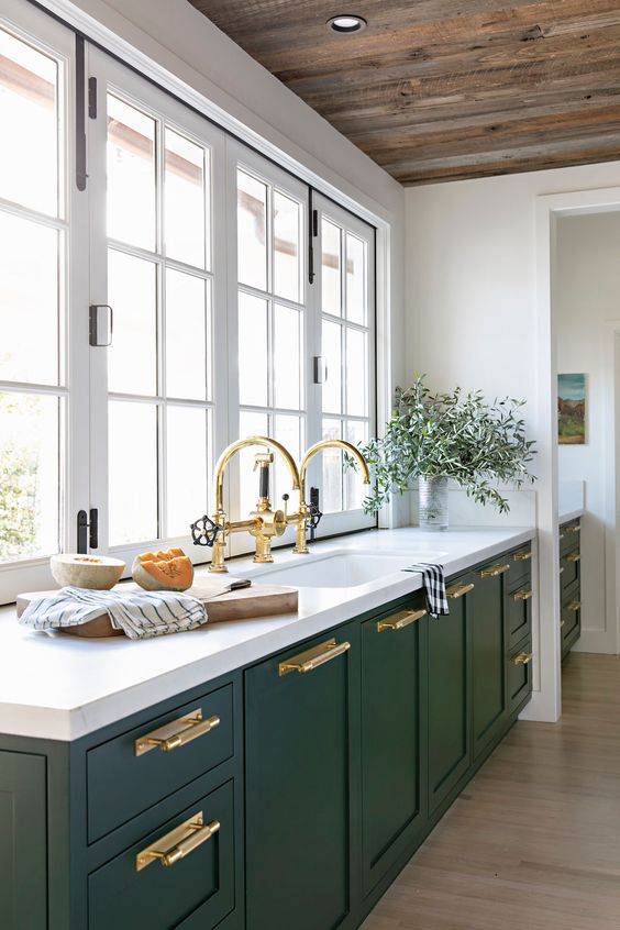 a hunter green modern kitchen with white stone countertops, gold handles and fixtures and a window instead of a backsplash