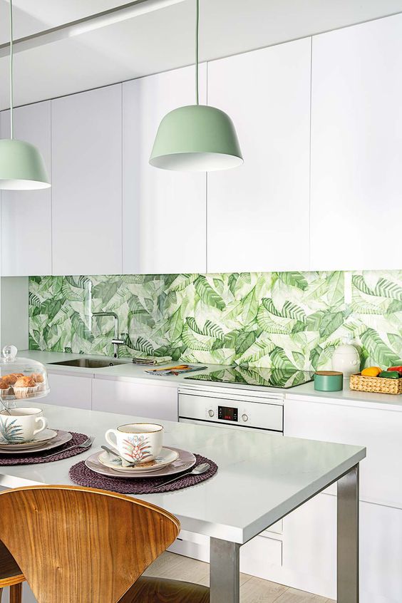 a minimalist white kitchen with a tropical leaf backsplash, green pendant lamps and touches of green here and there