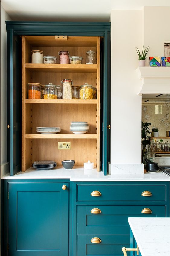 A chic teal kitchen with elegant cabinetry, light stained wooden compartments, gold fixtures and white stone countertops