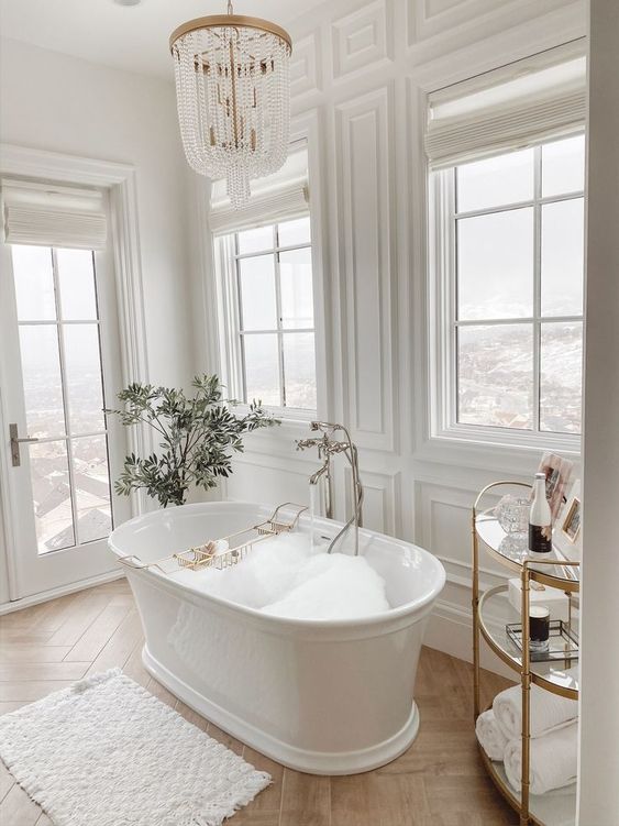25 a luxurious white bathroom clad with panels, with a gorgeous view, a vintage tub, a gold and glass side table and a crystal chandelier