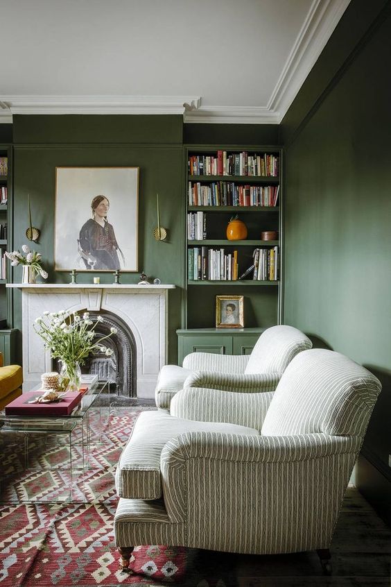 A sophisticated green living room with dark walls, vintage and modern furniture, built in bookshelves and a fireplace