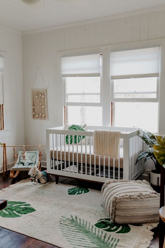 A neutral boho nursery with white and light stained furniture, tropical prints, a boho ottoman and macrame