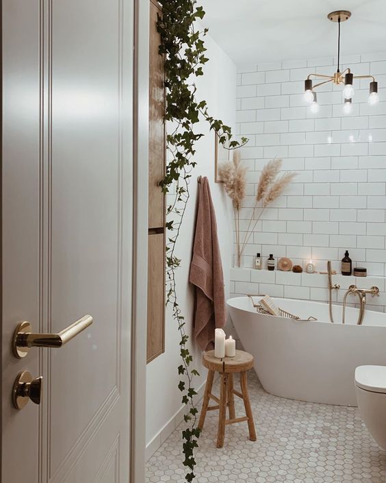 a pretty boho bathroom in white, with subway tiles, marble hex ones, an irregular tub and potted plants and a chandelier