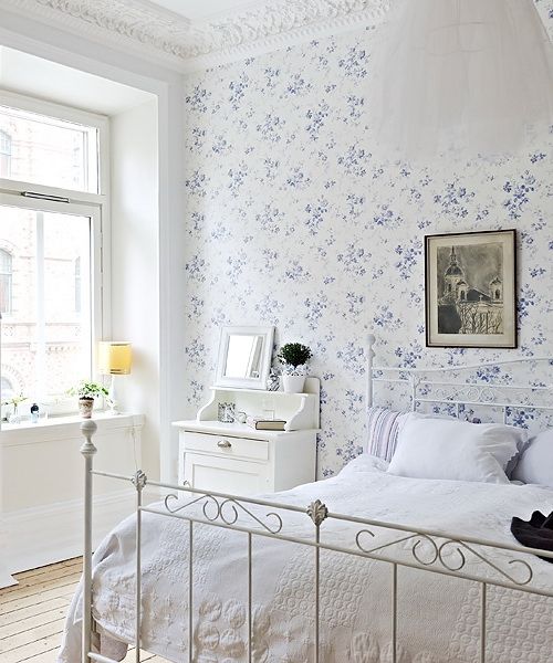 A vintage inspired bedroom with a blue floral wall, a white forged bed, white furniture, a dark artwork and a pendant lamp