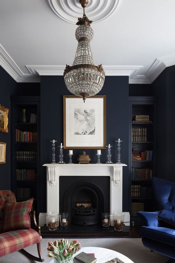 a moody living room with midnight blue walls, navy and plaid red furniture, a fireplace, a crystal chandelier and lots of books
