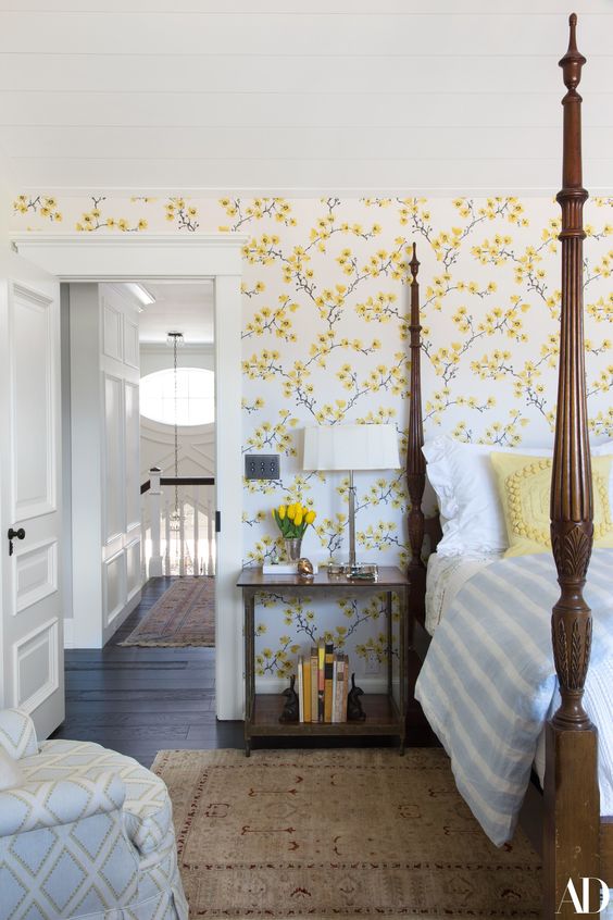 A vintage inspired bedroom with yellow floral walls, a heavy bed with pillars, a chair and nightstands plus blooms