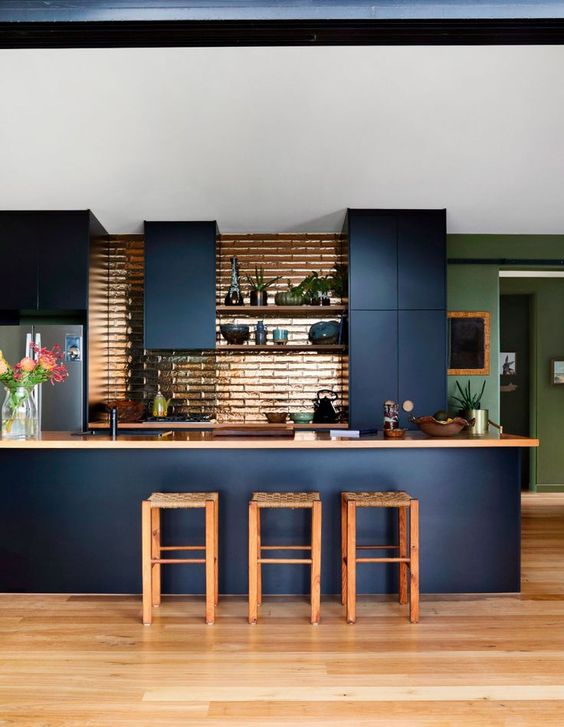 a chic and beautiful navy kitchen with sleek cabinets, tan countertops and a shiny copper tile backsplash