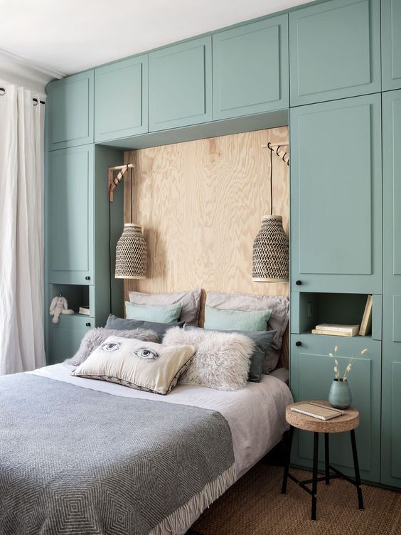 a chic small bedroom with a whole wall taken by a green storage unit, a plywood wall, sconces and cork stools is very cool