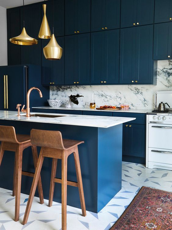 a glam navy ktichen with a white stone backsplash and countertops, a matching kitchen island and gold pendant lamps