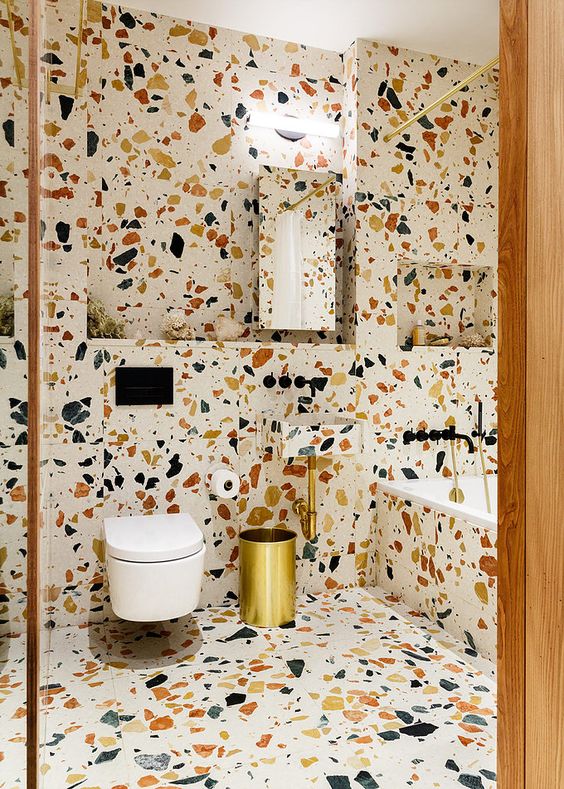 a bright terrazzo bathroom done with gold fixtures and a mirror plus a wall sconce over it