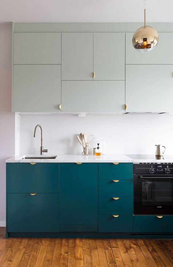 a stylish two tone kitchen in light green and teal, with gold handles, a copper pendant lamp and a white countertop and backsplash