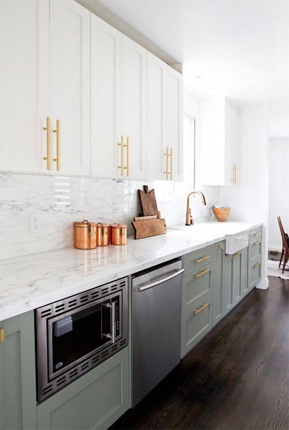 an elegant and shiny kitchen with white and sage green cabinetry, a white stone countertop and backsplash, gold touches