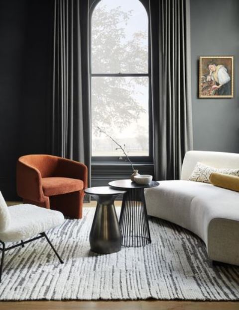 an exquisite and chic living room with grey walls, dark draperies, a curved sofa, a rust chair and a white one