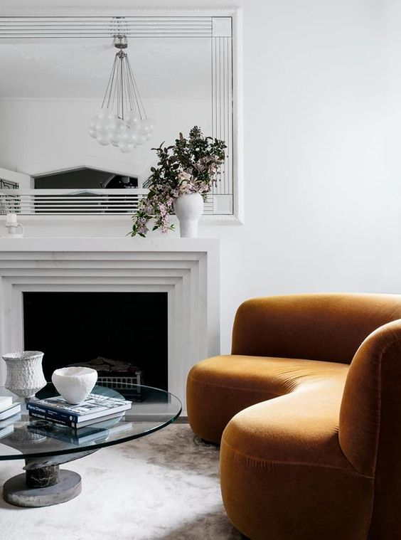 A refined living room with a fireplace, a rust colored curved sofa, a mirror, a round glass table