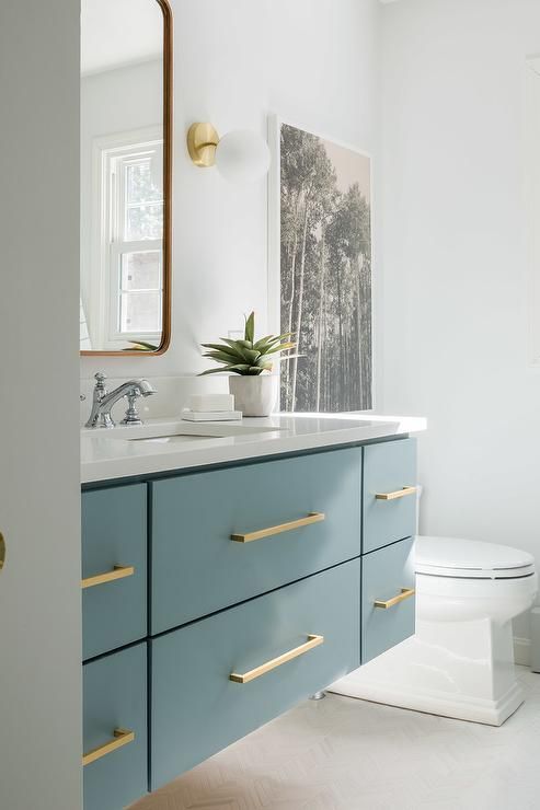 a small bathroom in neutrals, with a floating blue vanity, gold touches and a mirror with curved corners
